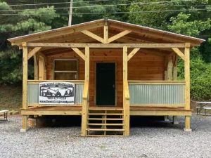 Front view of a small wooden cabin on a raised porch with steps a green door and a banner on the railing reading The West Virginia in a gravel lot with trees behind