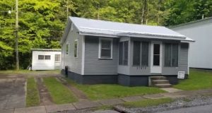 Small gray single story house with a covered front porch and a detached white garage to the left in a grassy yard with trees behind