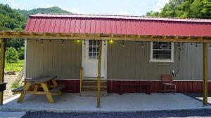 Small outdoor shelter with a red metal roof gray siding white door and string lights featuring a picnic table and bench on a concrete pad