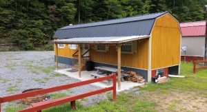 Small yellow cabin with a black foundation and metal roof featuring a covered porch a bench and a wood stack set on a gravel area with a forest backdrop