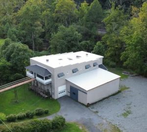 White multi section modern building with flat roof skylights and blue door set among dense trees with a gravel yard nearby